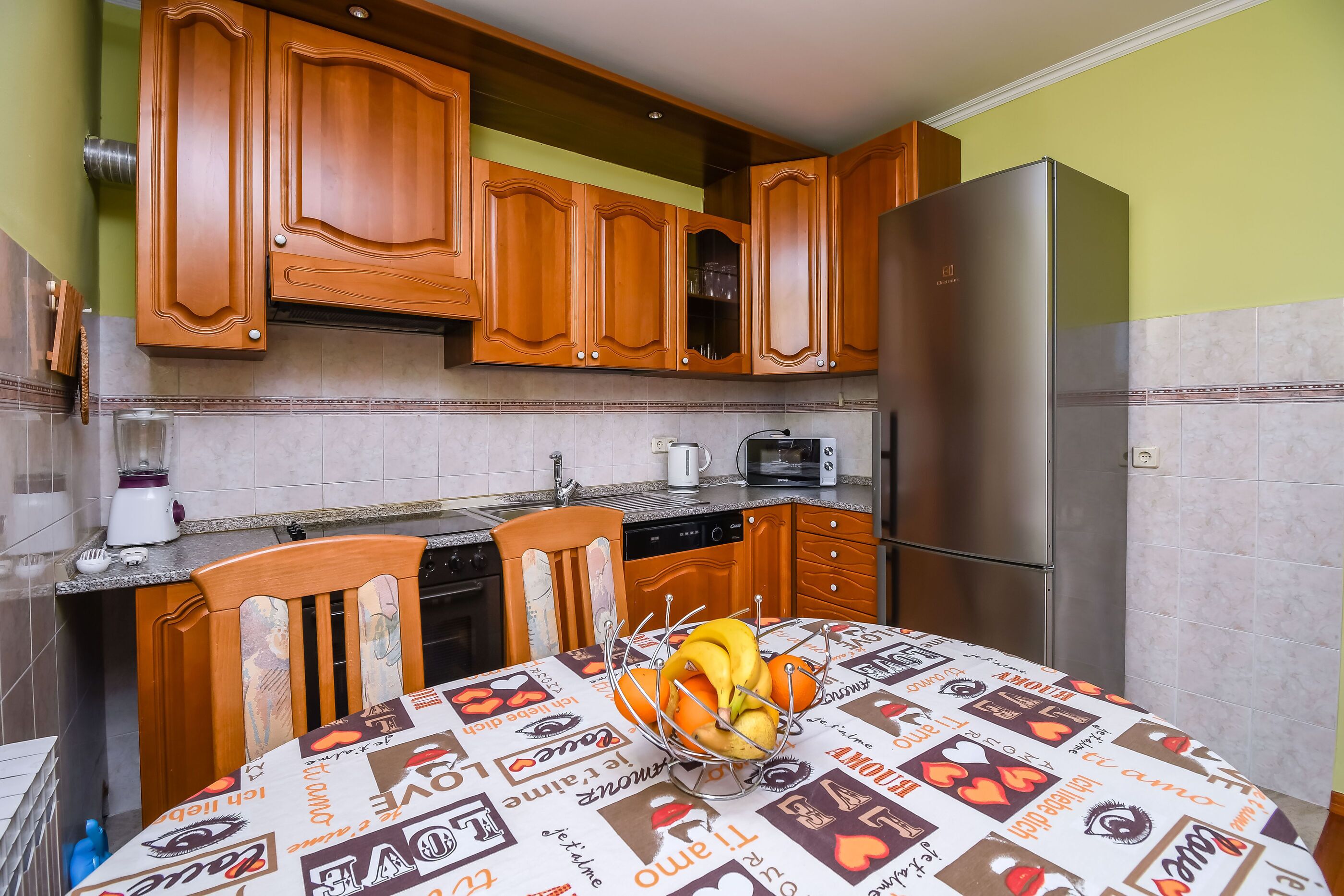 A kitchen with wooden cabinets, a stainless steel refrigerator, a dining table with a fruit bowl, and a coffee maker.