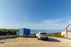 A white car parked near a blue covered trailer on a gravel driveway with a stone wall and a view of the sea and hills in the background. - Apartments Mrvelj (Jelsa)
