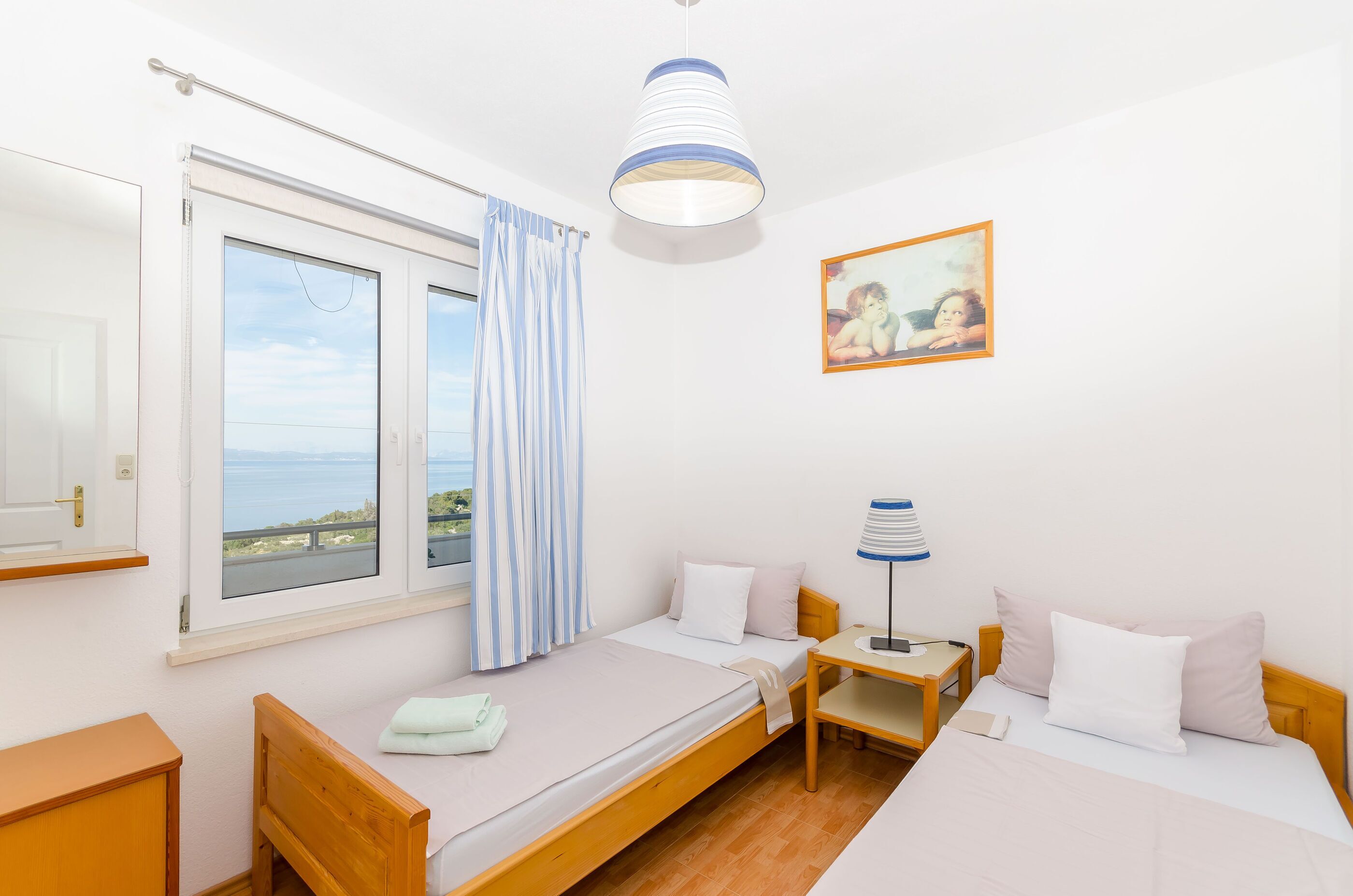 A bedroom with a bed, a desk, a lamp, a painting, and a window with a view of the sea.