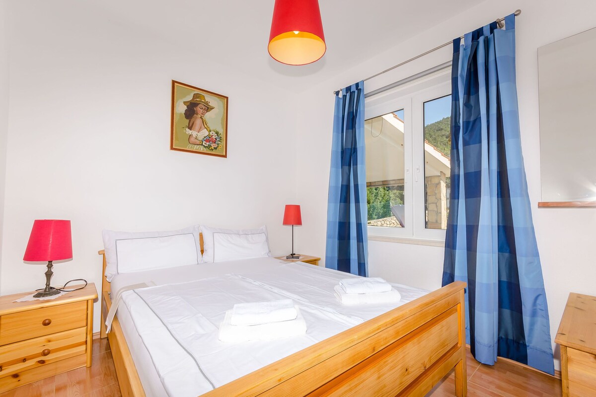 Una cama bien tendida con sábanas blancas, dos mesitas de noche con lámparas, una ventana con cortinas azules y un cuadro colgado en la pared.