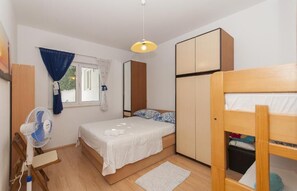 Une chambre à coucher de taille modeste, comprenant un lit simple, une armoire en bois, un bureau avec une chaise et un ventilateur sur pied.