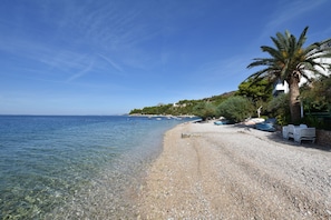 A pebble beach with clear water, a palm tree, and a row of white chairs. - House Fjaka (Omis)