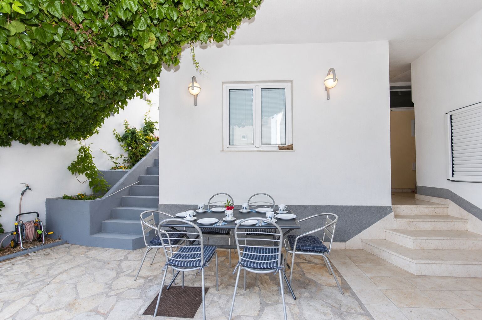An outdoor dining area with a table set for four, a staircase, and a wall-mounted light fixture.
