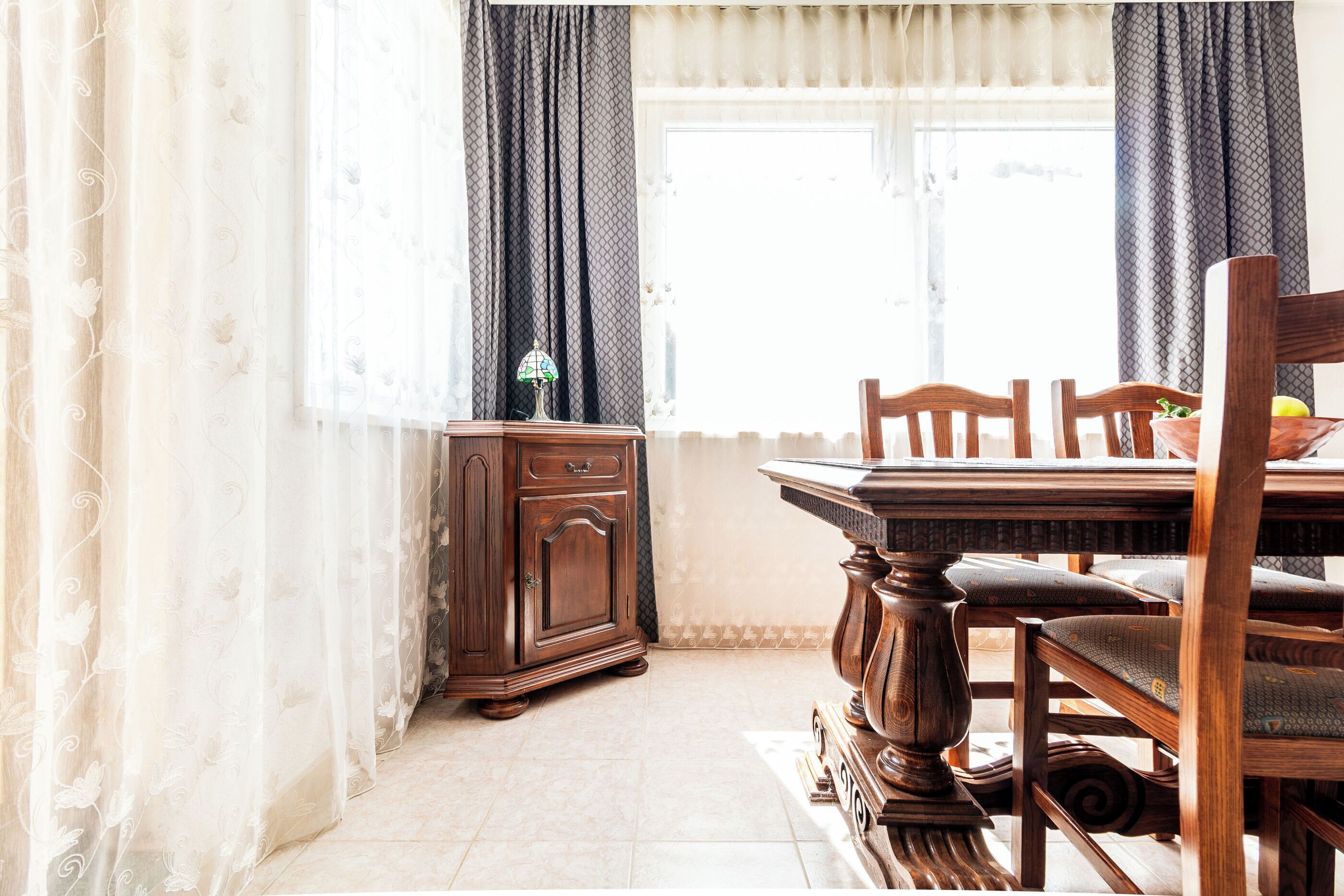 Een eethoek met een houten tafel en stoelen, een kast, en een raam met decoratieve gordijnen.