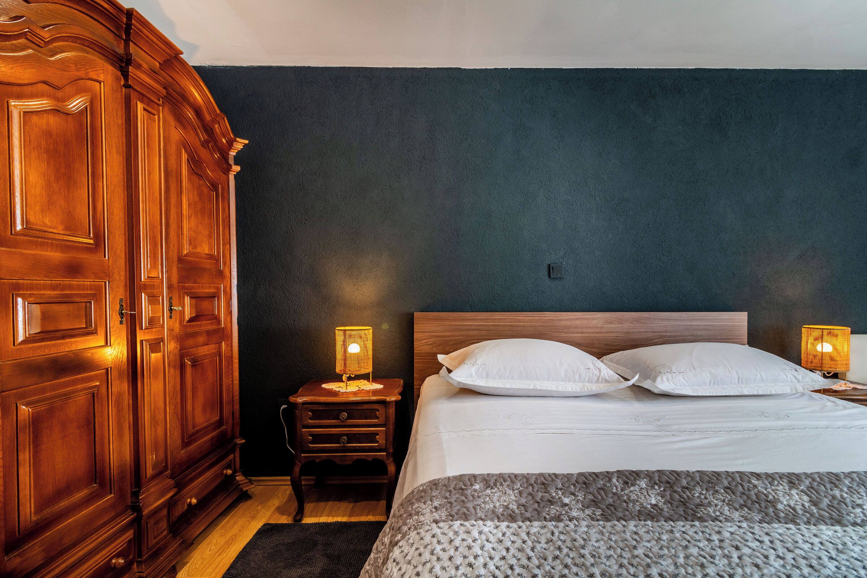 Un dormitorio con un armario de madera grande, una mesita de noche con lámpara y una cama con ropa blanca.