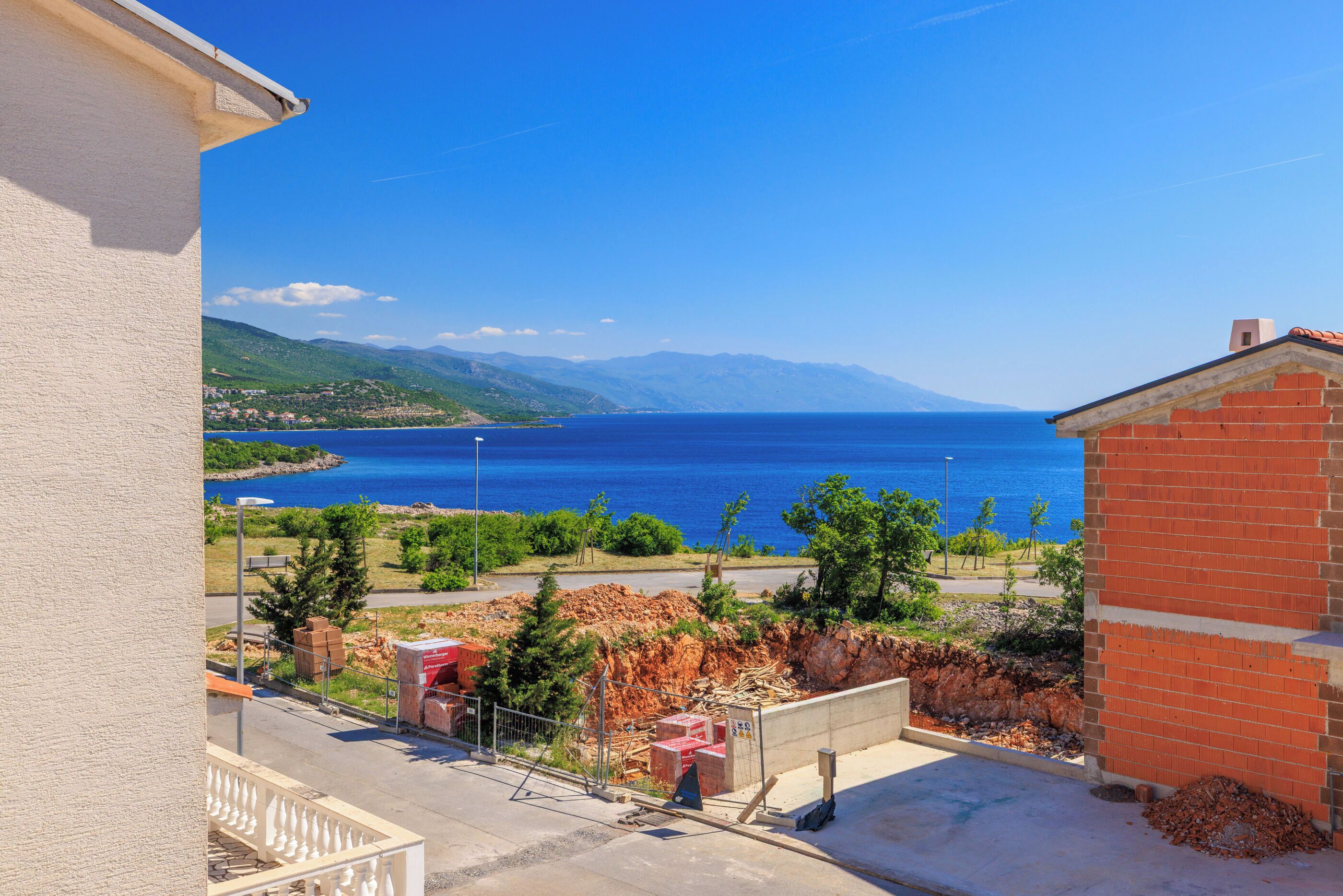 Blick vom Balkon auf eine Küstenlandschaft mit blauem Meer, entfernten Bergen und einer kleinen Baustelle.