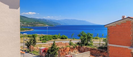 Blick vom Balkon auf eine Küstenlandschaft mit blauem Meer, entfernten Bergen und einer kleinen Baustelle.