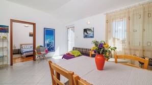 Un vaste espace de vie comprenant une table à manger, des chaises, un canapé et un lit. Une grande fenêtre est agrémentée de rideaux, un tableau orne le mur et un vase rempli de fleurs est posé sur la table.