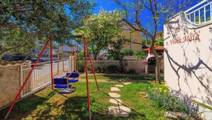 Une villa avec un chemin de pierre menant à une aire de jeux avec des balançoires, entourée de verdure et d’un mur de pierre.