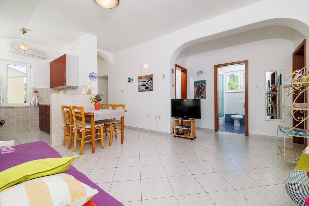 A spacious living area with a dining table, chairs, a TV, and a kitchen in the background.