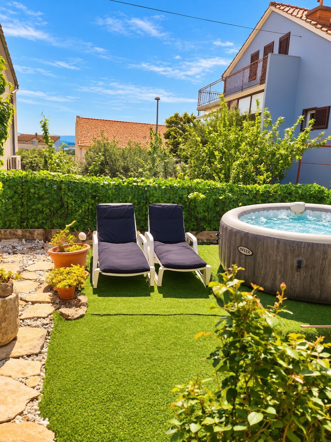 A backyard with artificial grass, two lounge chairs, a hot tub, and a house with a red-tiled roof.