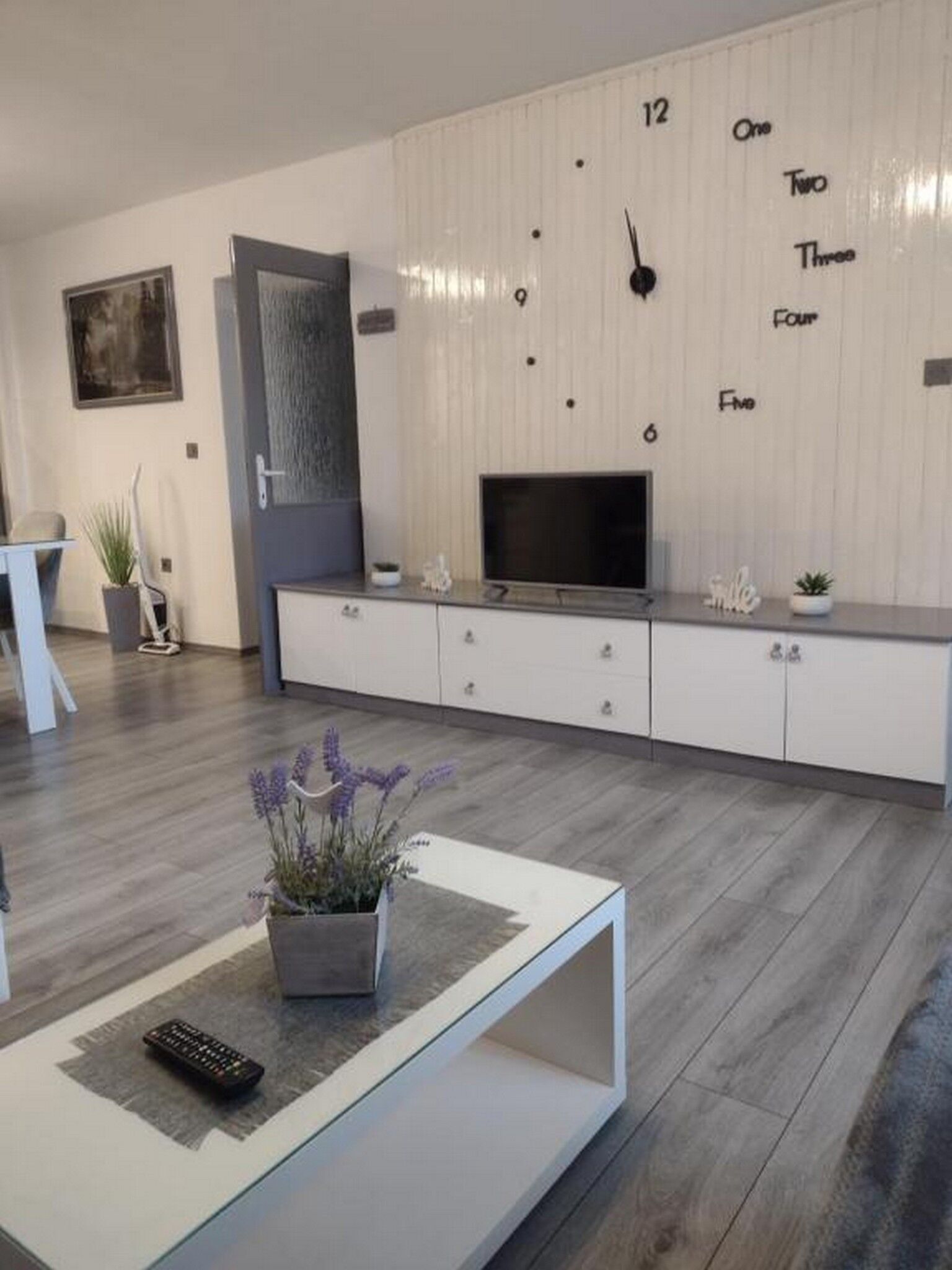 A modern living room with a flat-screen TV, a coffee table with a remote, and a clock on the wall.