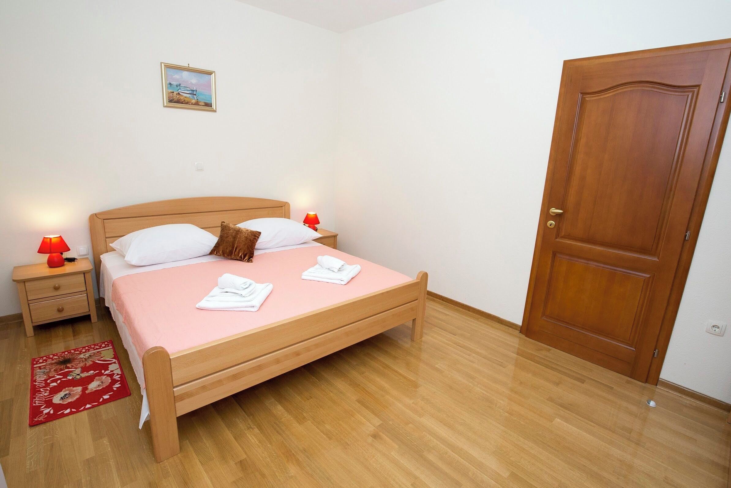 Un dormitorio con una cama de madera, dos mesitas de noche con lámparas, una puerta y un pequeño cuadro en la pared.