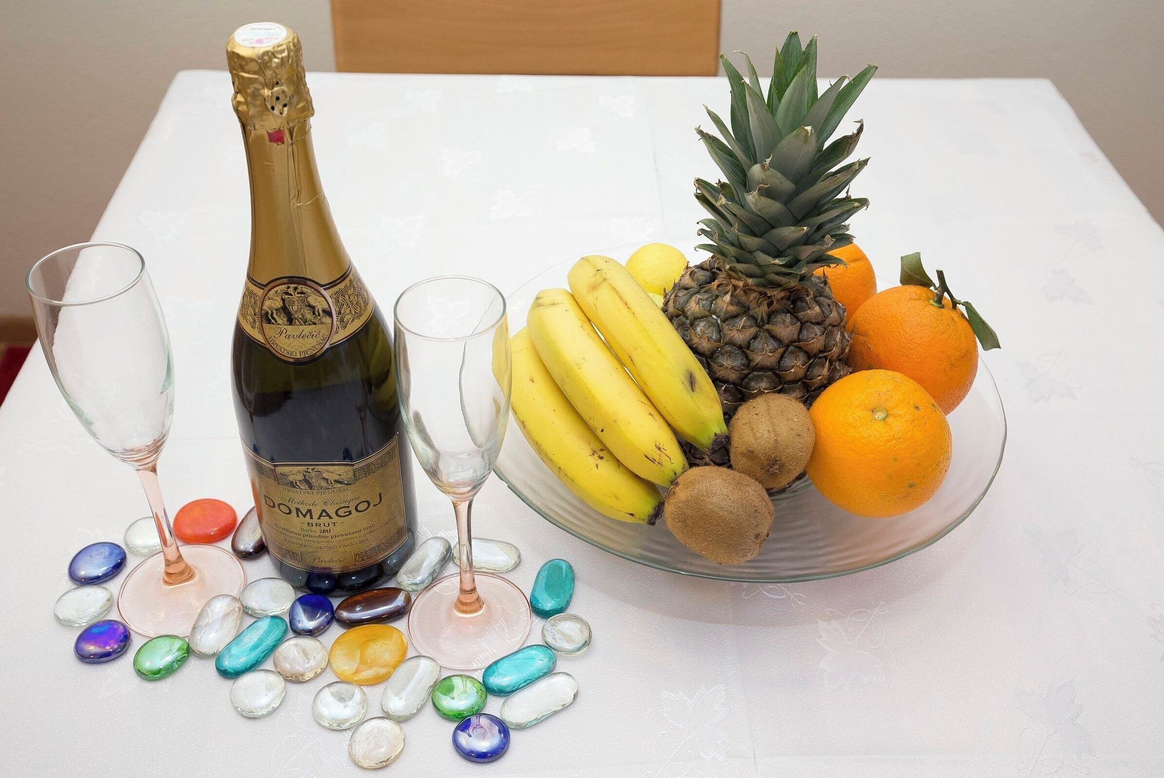A bottle of Domagoj Brut champagne, two glasses, a bowl of fruit, and colorful stones on a table.
