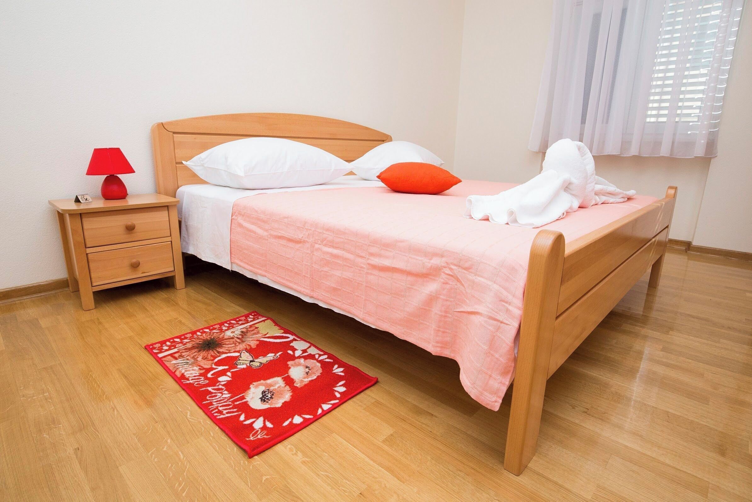 A neatly made bed with a pink bedspread and white pillows, a wooden nightstand with a red lamp, and a red floral rug on a wooden floor.