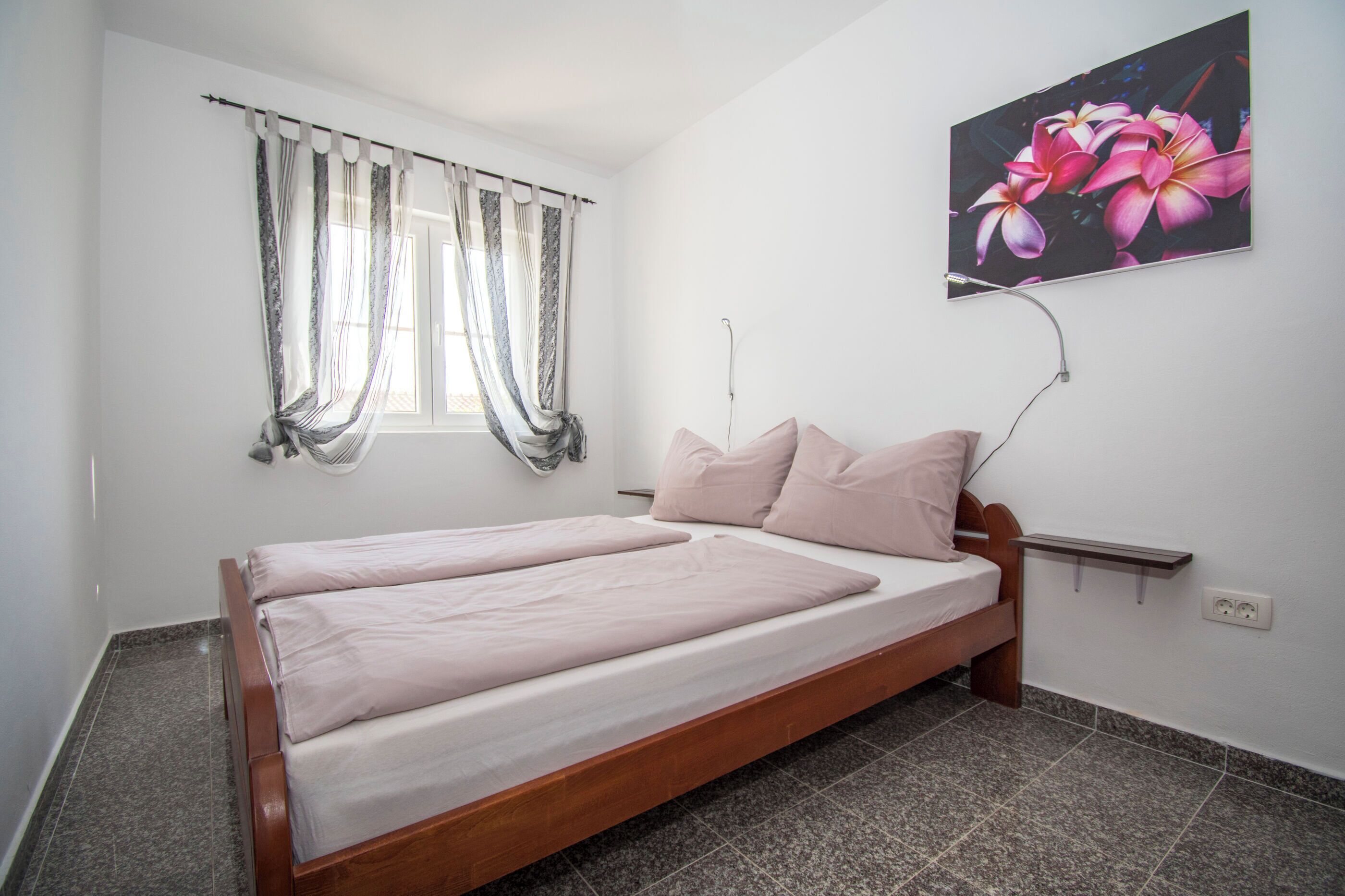 A single bed with a wooden frame and a headboard, a window with curtains, a wall-mounted power outlet, and a framed picture of flowers.