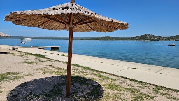 Ein Strandsonnenschirm mit runder Basis, ein Betonweg und ein stiller See mit Booten im Hintergrund.