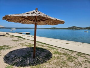 A beach umbrella with a circular base, a concrete walkway, and a calm lake with boats in the background. - Apartments Maslina (Bilice)