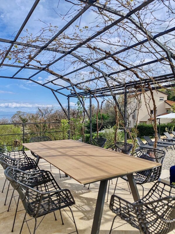 An outdoor patio with a wooden table and black metal chairs, surrounded by a metal pergola structure. - Apartment Aria (Lovran)