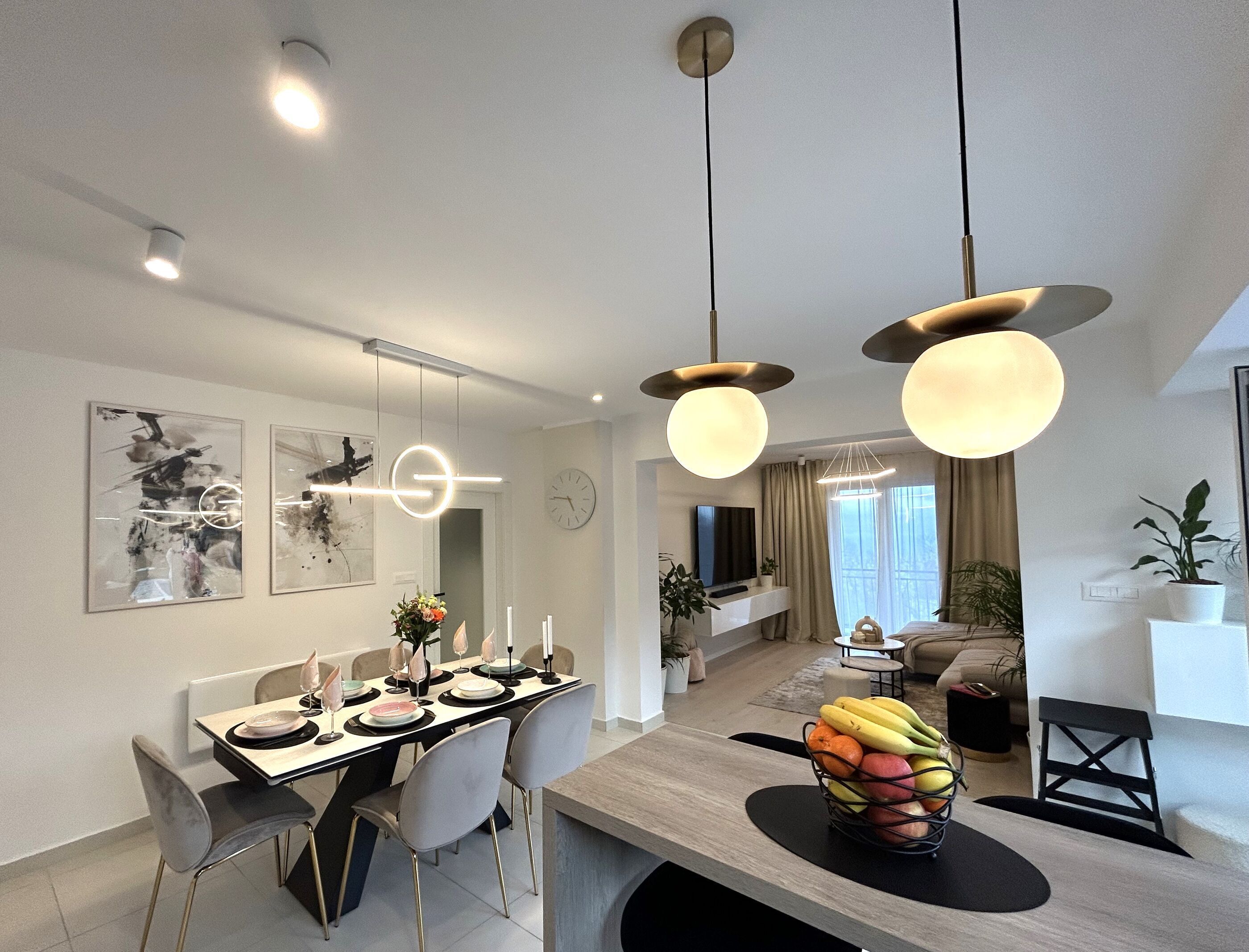 A modern dining area with a wooden table, grey chairs, and a fruit bowl. There are ceiling lights and a flat-screen TV in the background.