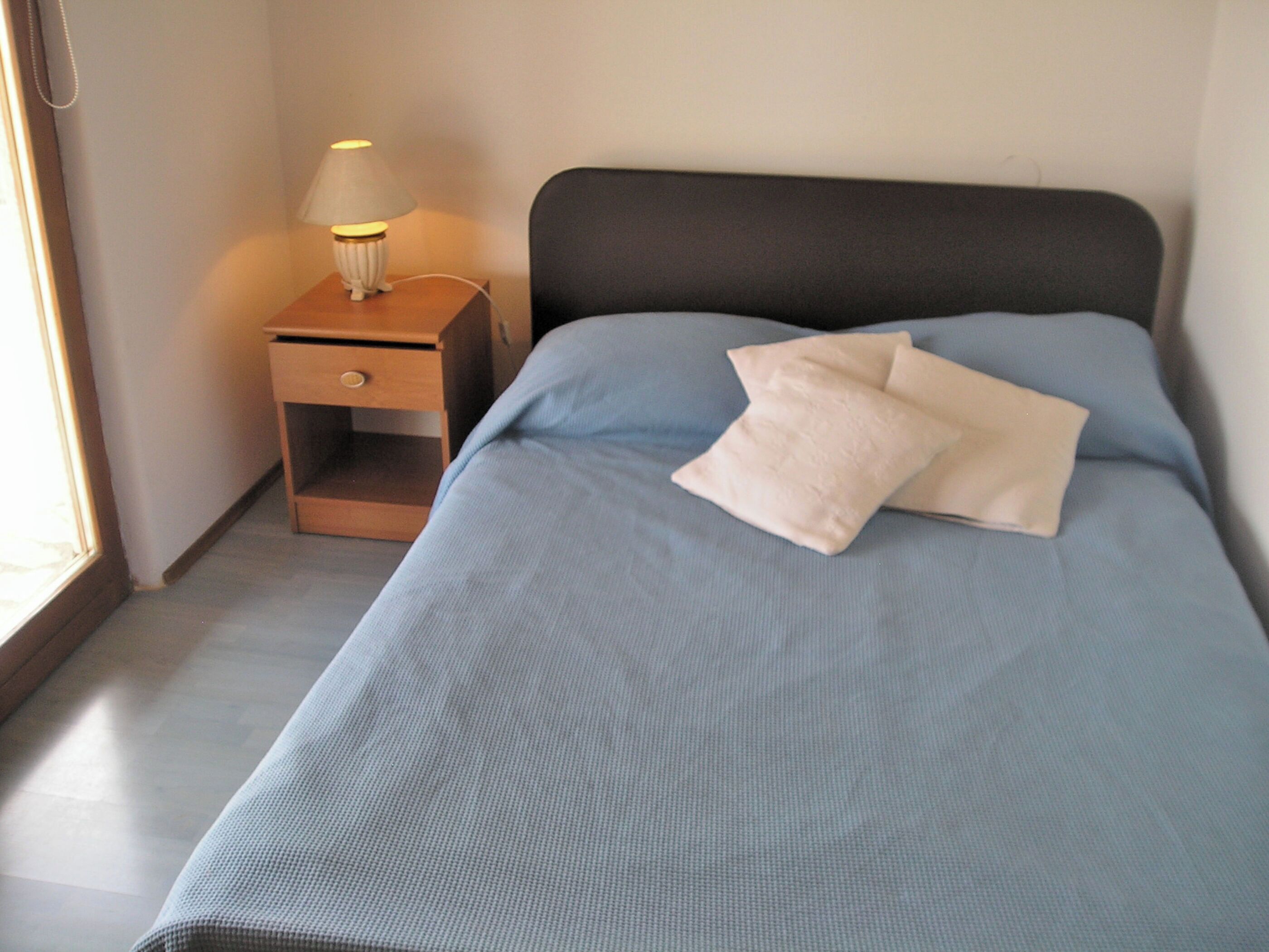 A neatly made bed with a blue bedspread and two pillows, a small wooden nightstand with a lamp, and a window to the left.