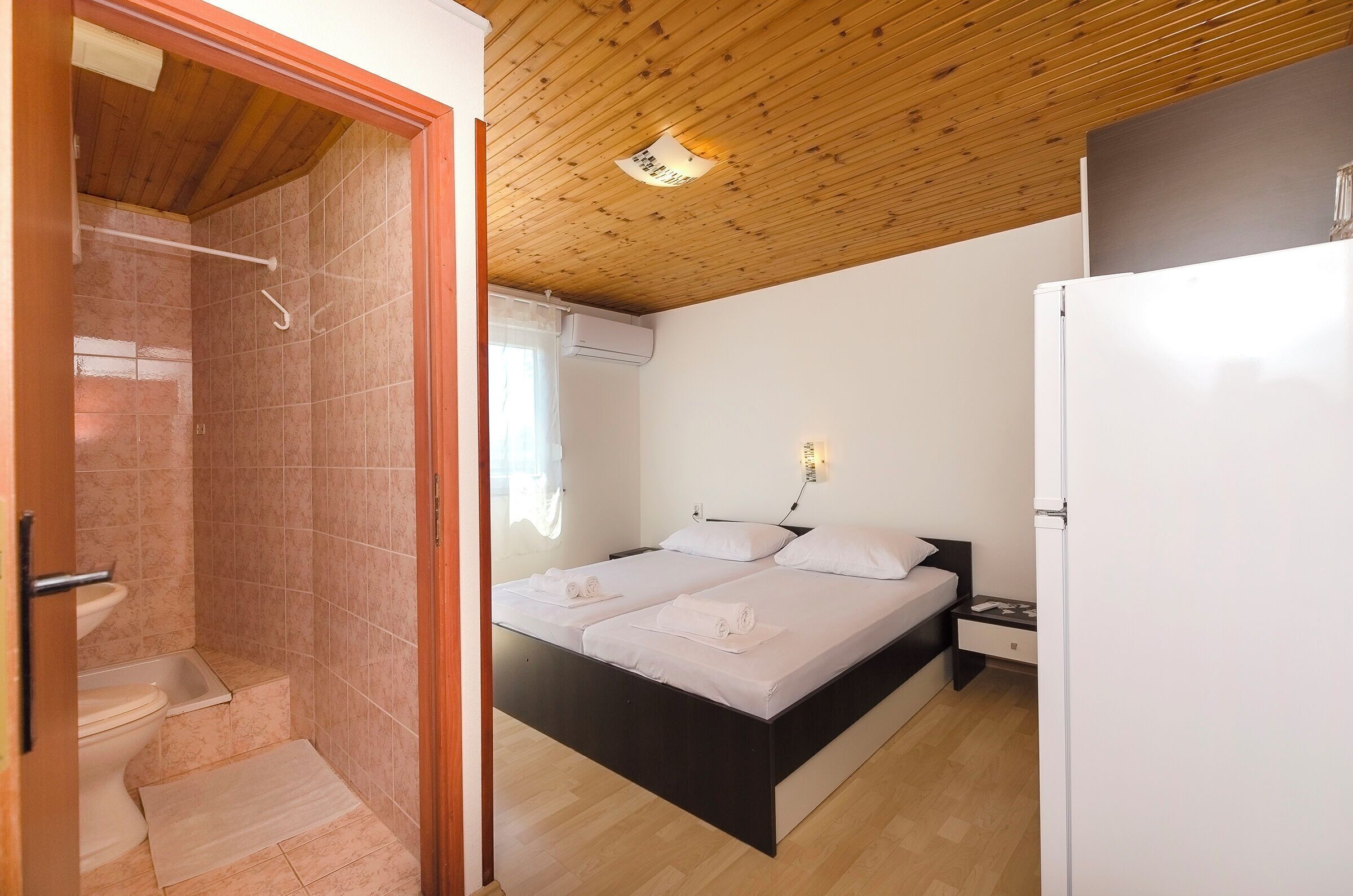 A bedroom with a wooden ceiling, a bed with white linens, a nightstand, and a white refrigerator.