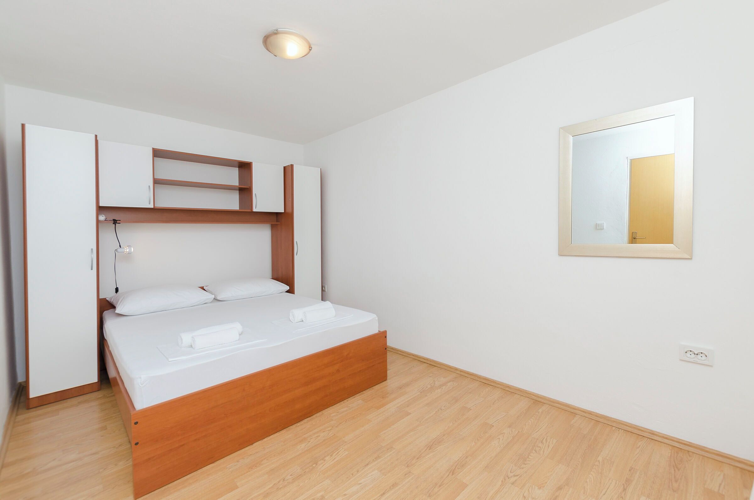 A bedroom with a bed, wooden headboard, built-in shelving, a bedside table, and a mirror on the wall.