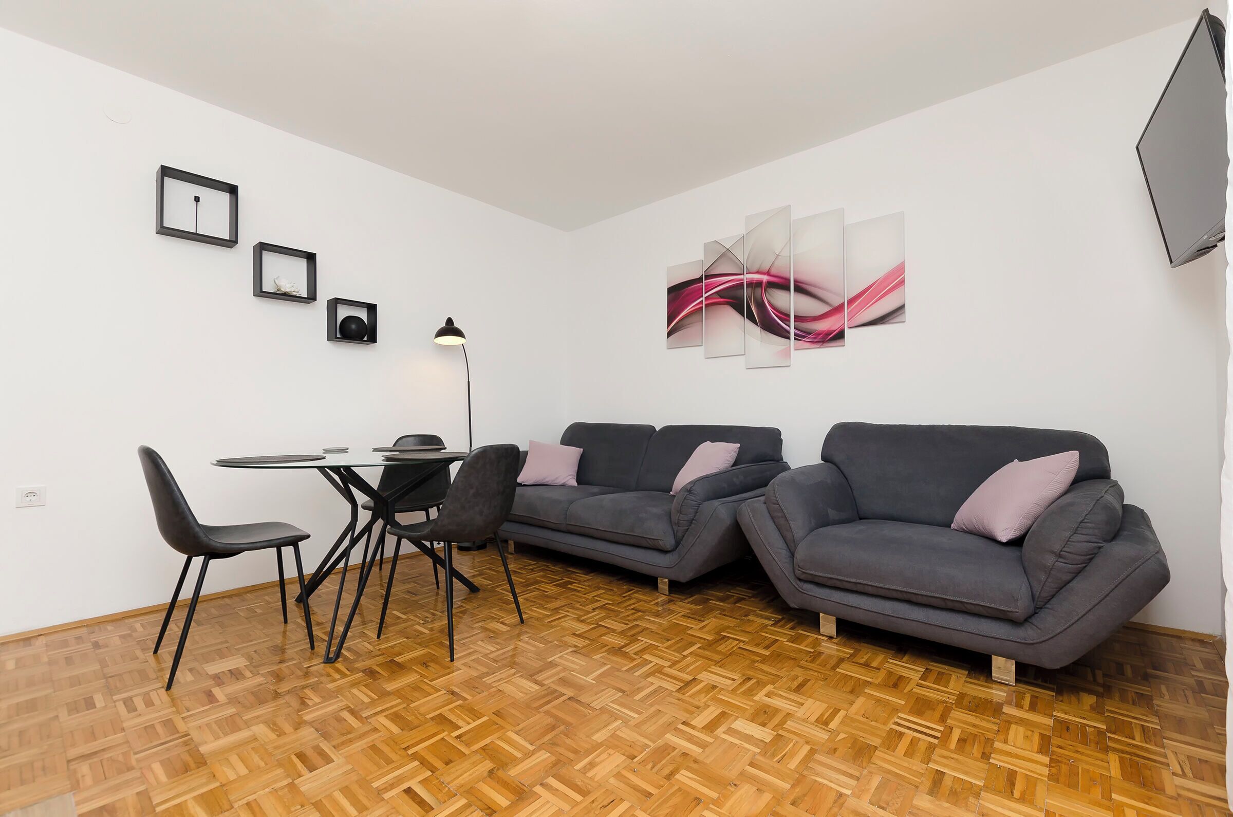 Une pièce avec un parquet en bois, un canapé gris avec des coussins, une petite table avec des chaises et un téléviseur fixé au mur.