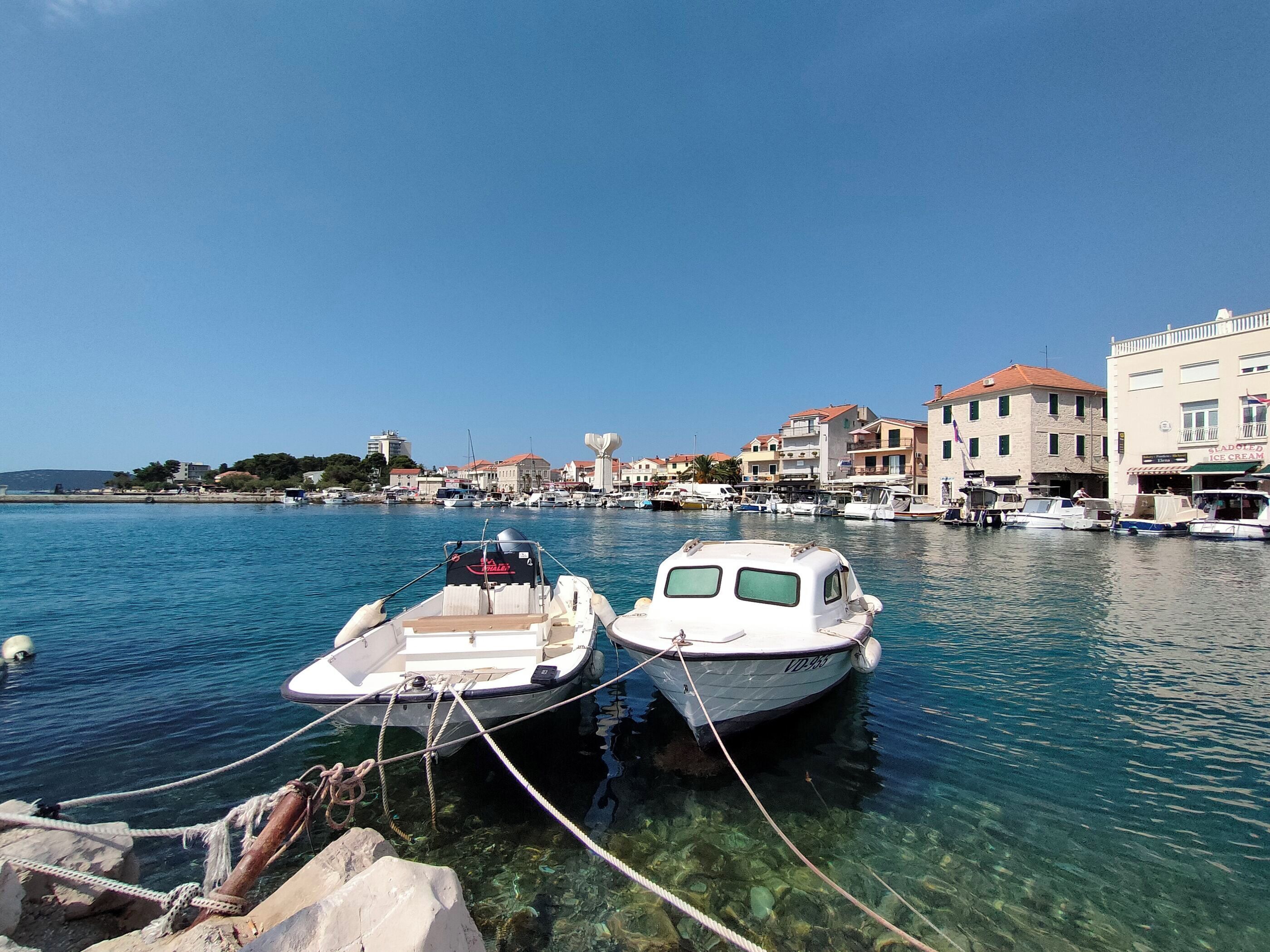 A marina with boats docked, clear water, and buildings in the background.