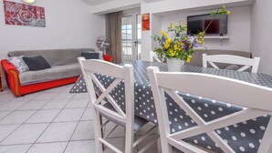 Un salon moderne avec un canapé gris, une table à manger entourée de chaises blanches, un téléviseur fixé au mur et une grande fenêtre.