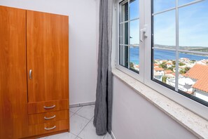 Quarto com guarda-roupa de madeira, janela com vista para o mar e edifícios, e cortina.