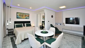 Moderni hotellihuone, jossa on suuri sänky, ruokapöytä tuoleineen, televisio ja taideteoksia seinillä.