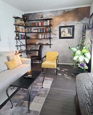 Una sala de estar con un sofá, un sillón amarillo, una mesa de centro, una estantería y una planta en maceta.