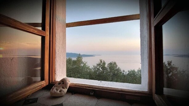 Ein Zimmer mit großem Fenster, Blick auf ein Gewässer und entfernte Landschaft.