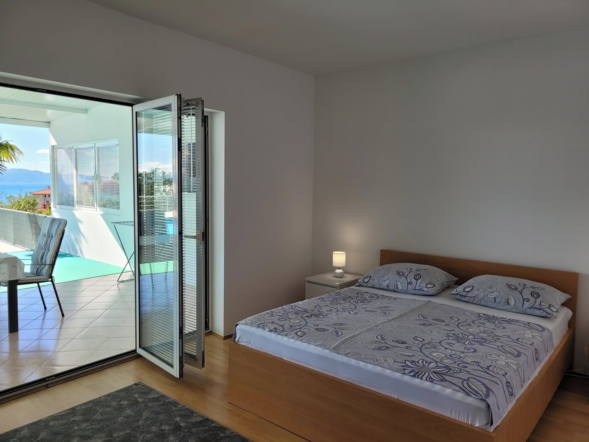 A bedroom with a bed, a nightstand, a lamp, and a sliding glass door leading to a balcony with a view of the sea.