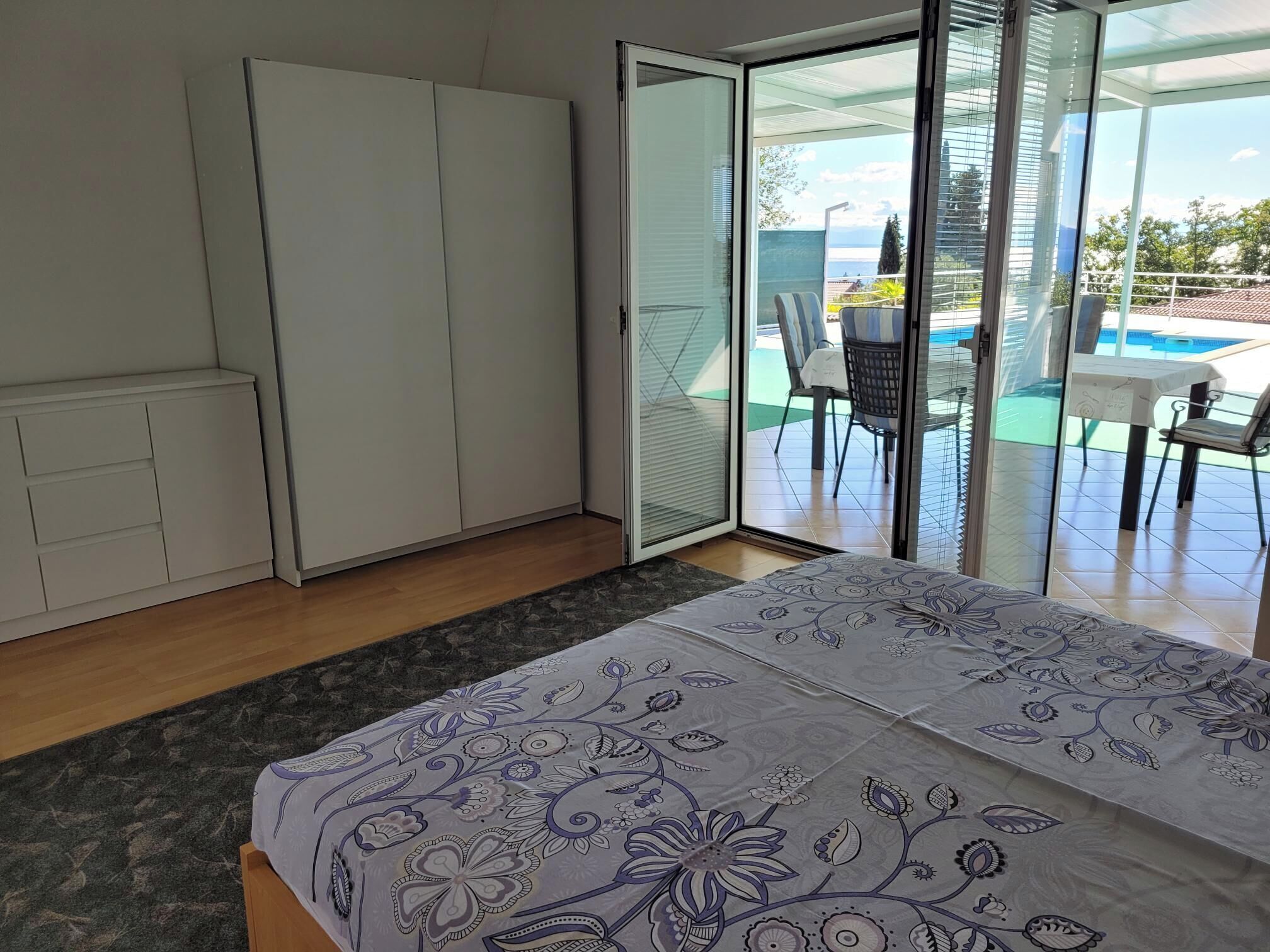 A bedroom with a bed, wardrobe, and a sliding glass door leading to a balcony with a table and chairs.