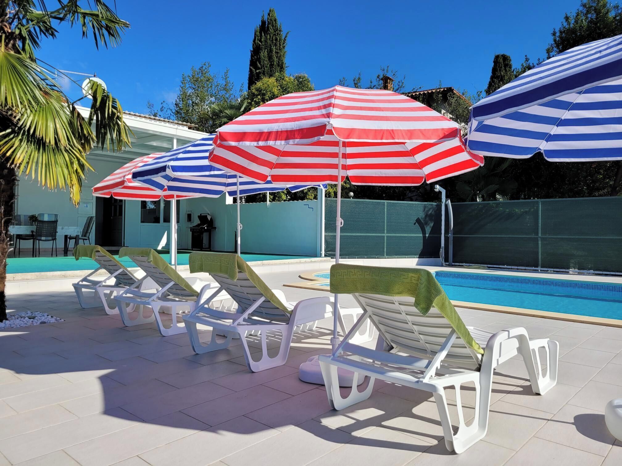 Area piscina con ombrelloni a righe, poltrone prendisole e una piscina blu cristallina.