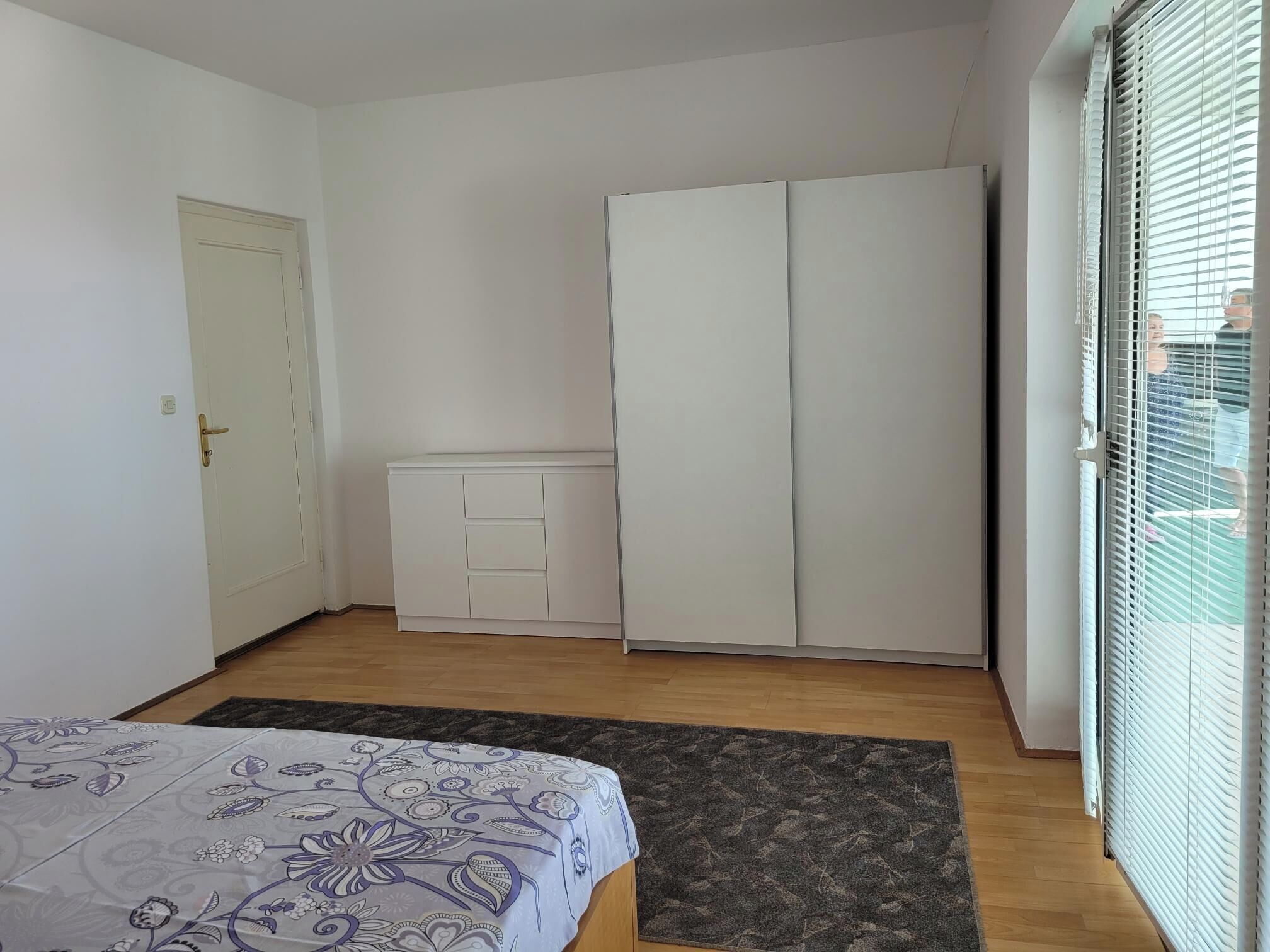 A bedroom with a bed, a wardrobe, a small cabinet, and a door.