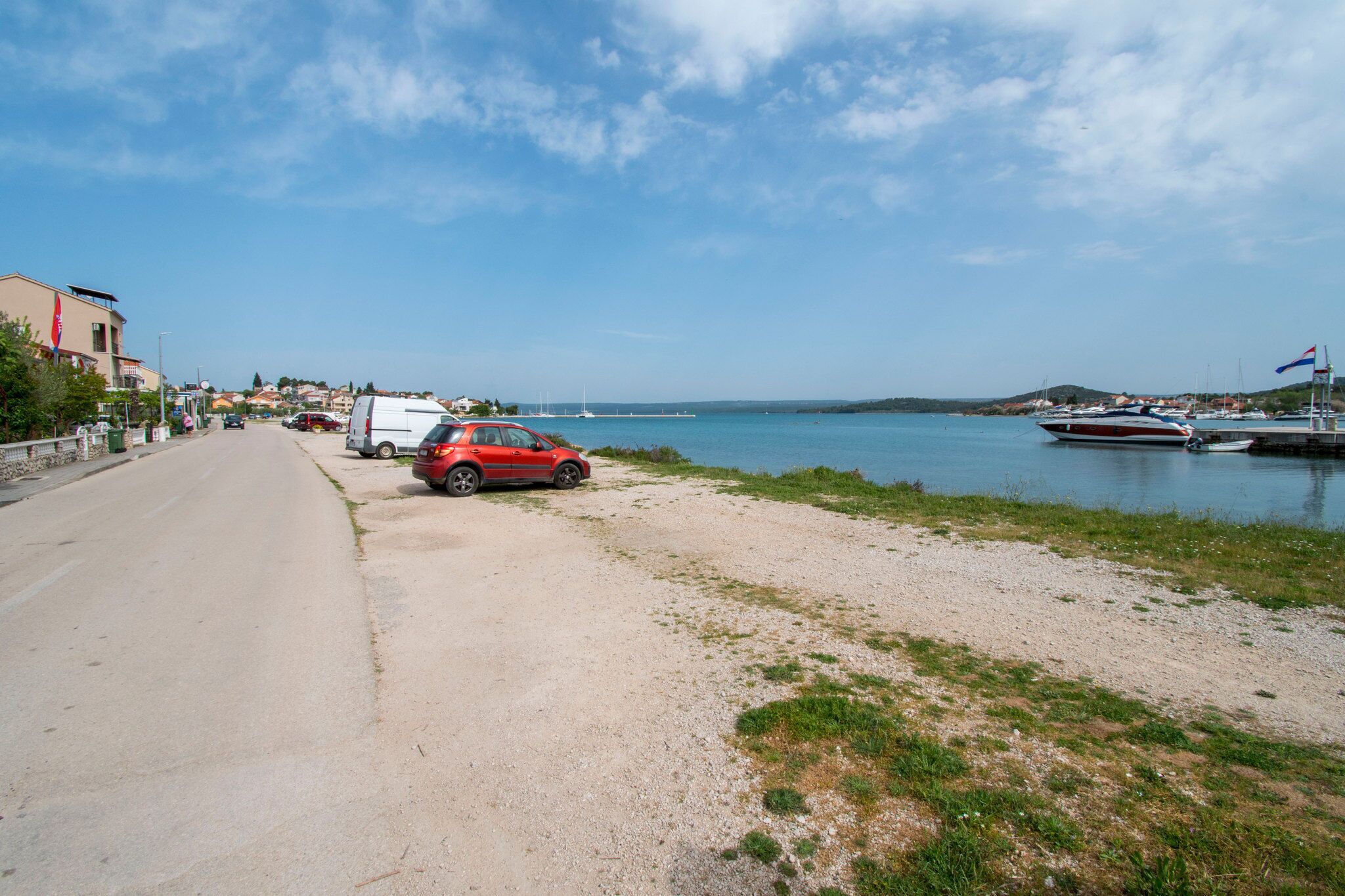 A coastal road with a red car parked on the side, a marina with boats, and buildings in the background.