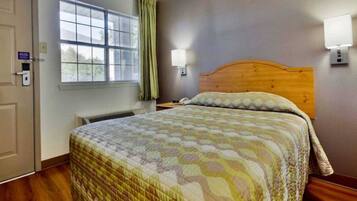 A hotel room with a bed, a window with curtains, a wall-mounted light, and a thermostat.