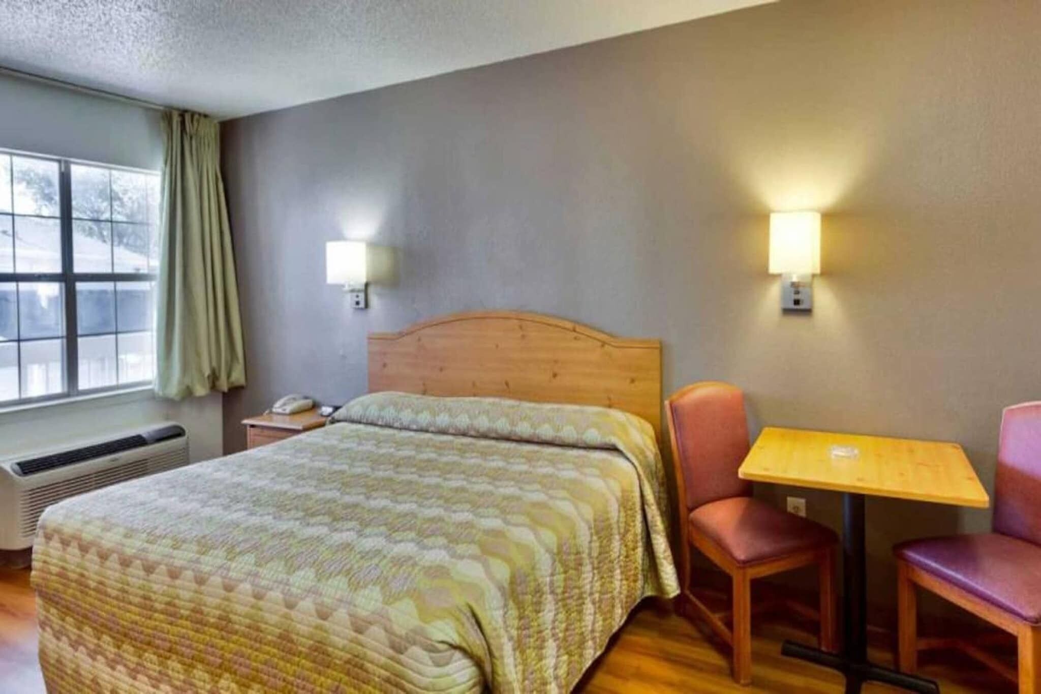 A hotel room with a bed, a table, two chairs, a nightstand, a lamp, and a window with curtains.
