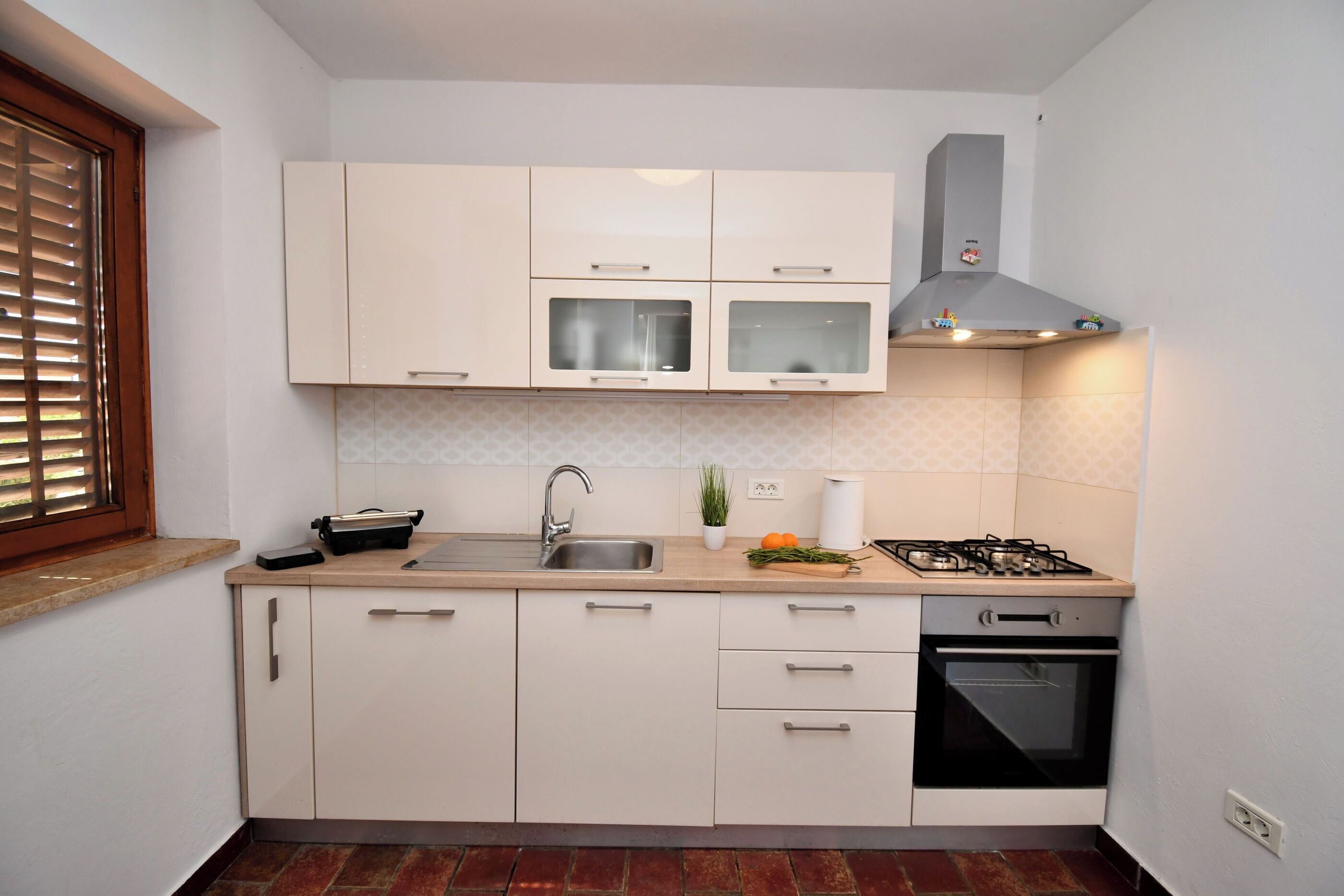 Una cocina compacta con armarios blancos, cocina a gas, horno, fregadero y una ventana con persianas.