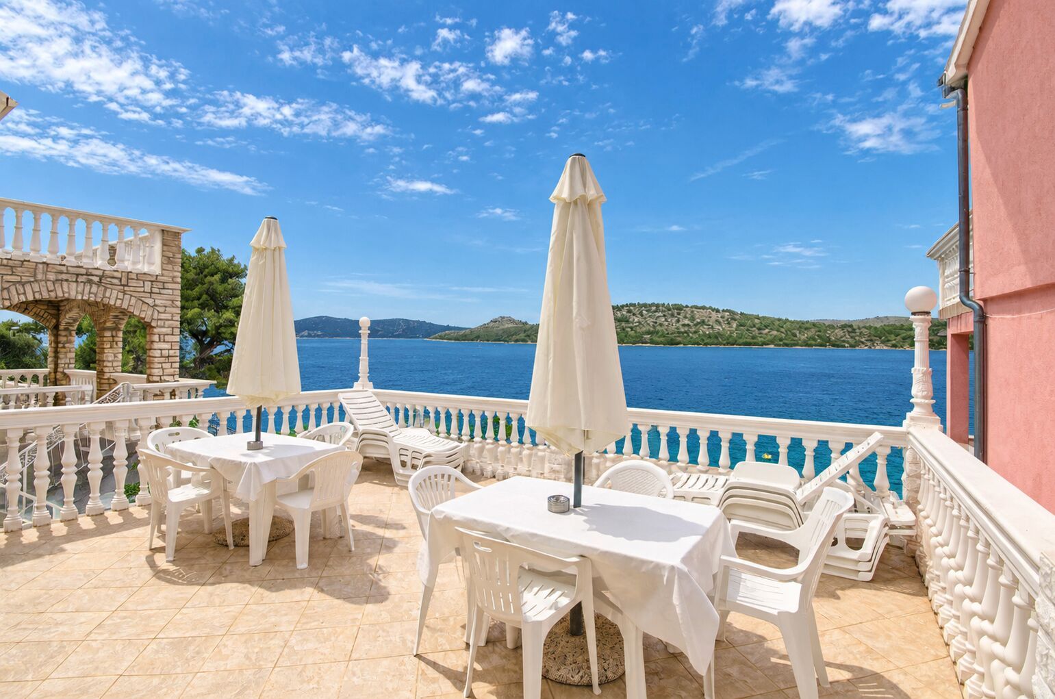 Eine Terrass mit weißen Tischen und Stühlen, Sonnenschirmen und Blick aufs Meer.
