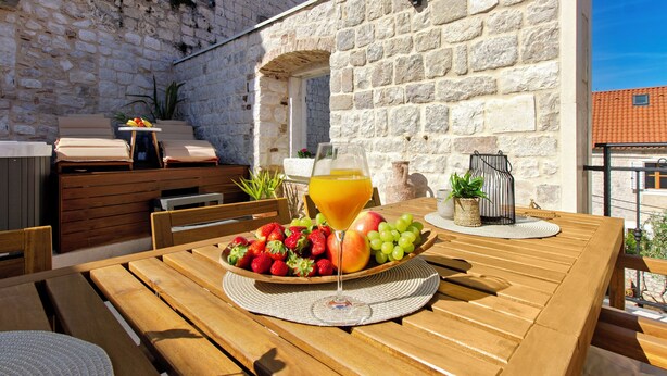 Une table en bois, sur laquelle se trouvent un panier de fruits et un verre de jus d’orange, est placée devant un mur de pierre agrémenté d’une petite arche.