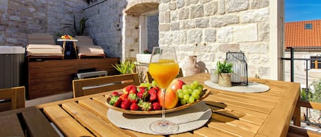 Une table en bois, sur laquelle se trouvent un panier de fruits et un verre de jus d’orange, est placée devant un mur de pierre agrémenté d’une petite arche.