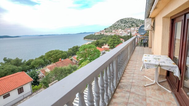 A balcony with a table and chairs, overlooking a coastal town and the sea. - Apartments Marinero (Pakostane)
