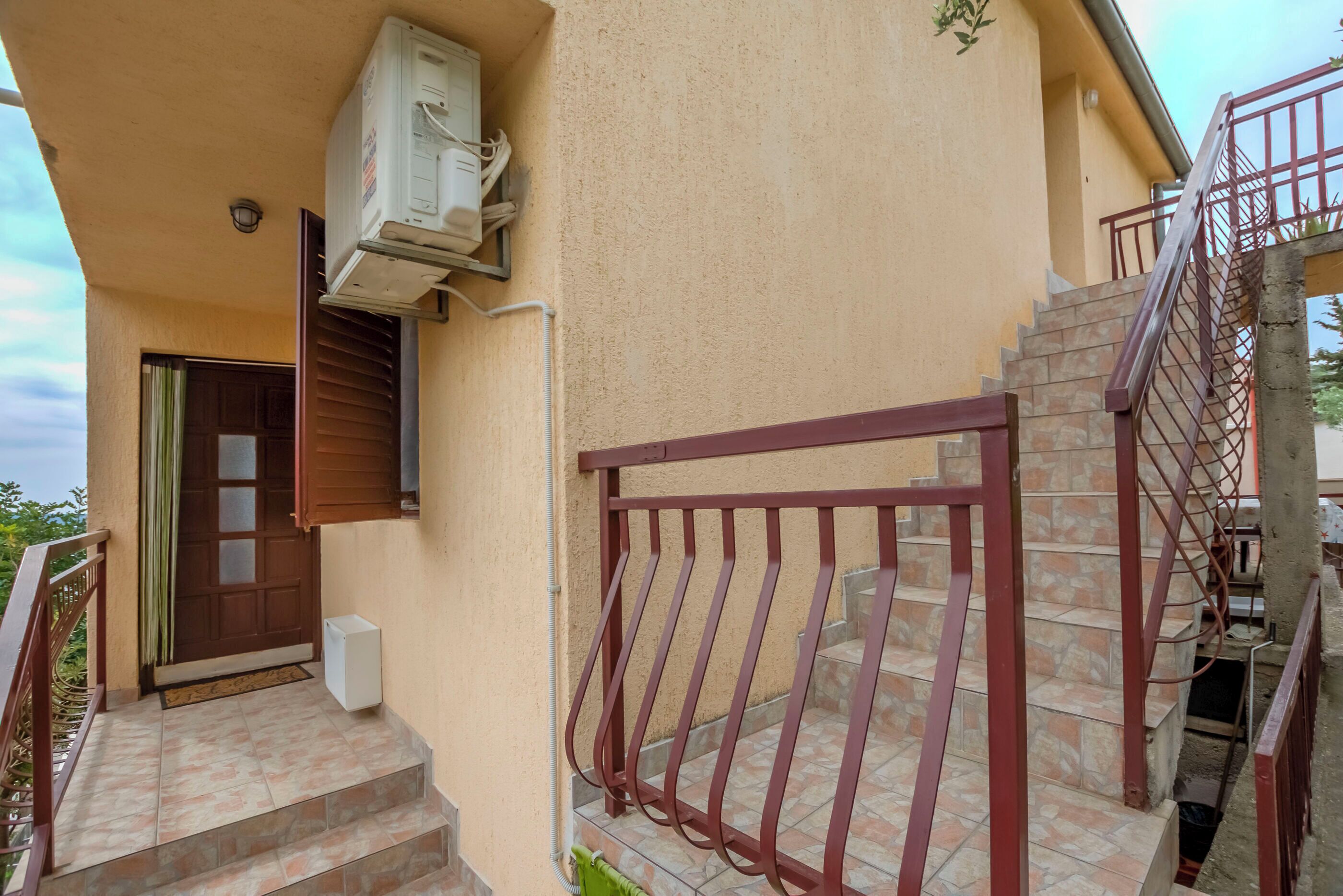 Un edificio con una puerta marrón, un pequeño equipo de aire acondicionado y una escalera con barandilla metálica.