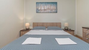 A neatly made bed with a geometric patterned bedspread, two pillows, and two folded towels. There are two bedside tables with lamps and a painting of sailboats on the wall above the bed. - Apartments Murter Jurjako (Murter-Kornati)