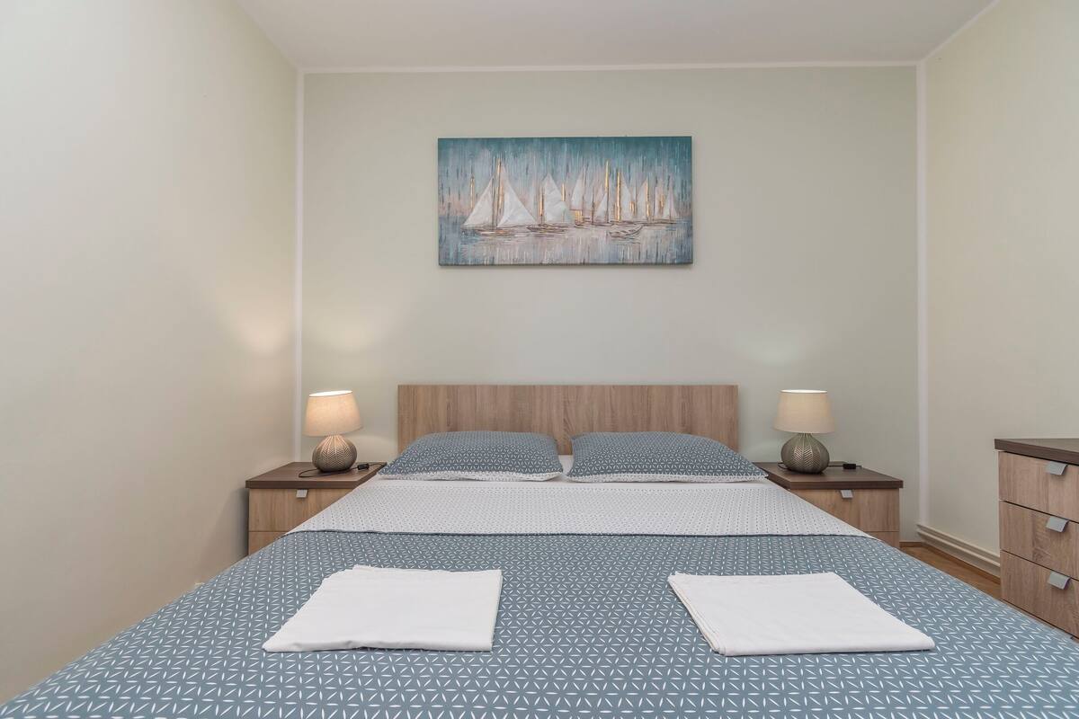 A neatly made bed with a geometric patterned bedspread, two pillows, and two folded towels. There are two bedside tables with lamps and a painting of sailboats on the wall above the bed.
