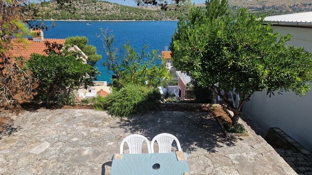 A stone patio with two white chairs and a striped table, overlooking a body of water and a hillside. - Apartment Cosy Terrace (Sibenik)
